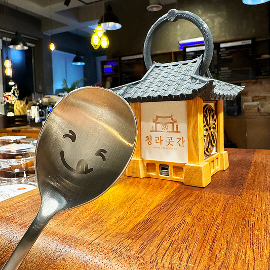 In front of a traditional Korean Hanok miniature, a spoon with a winking face is greeting you.