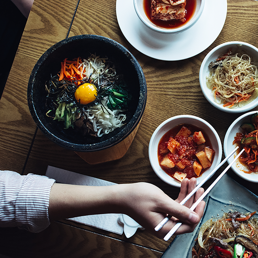 A hand holding flat chopsticks hovers over a table set with Korean dishes like kimchi, bibimbap, and seasoned vegetables.