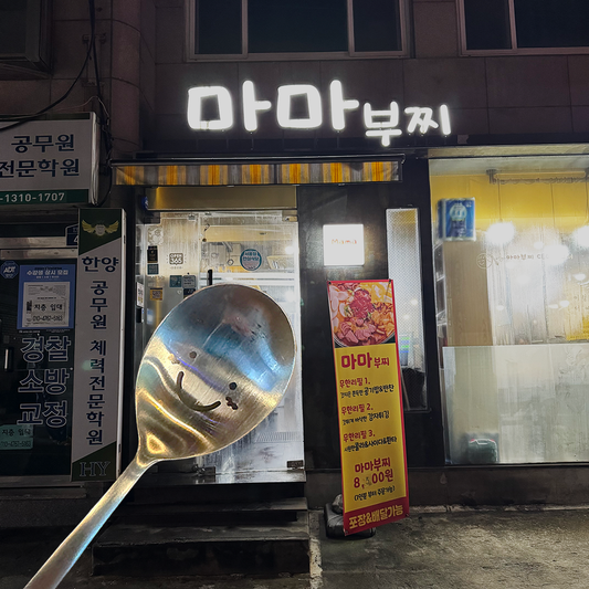 In front of the signboard that says 'Mama bujji' in Korean, there's a smiling spoon that warmly welcomes you.