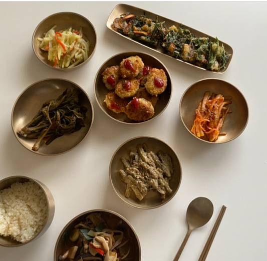 On a white table, a spoon and chopsticks are placed alongside stir-fried potatoes, kimchi, rice, soup, and various side dishes.